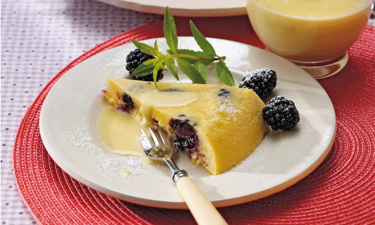 Fetta di omelette dorata con more scure incorporate, velata da salsa bianca cremosa, decorata con foglie verdi fresche su piatto chiaro