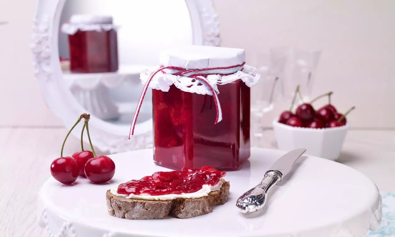 Confiture rouge brillante dans un pot décoré avec un morceau de pain tartiné et des cerises fraîches sur un plat blanc