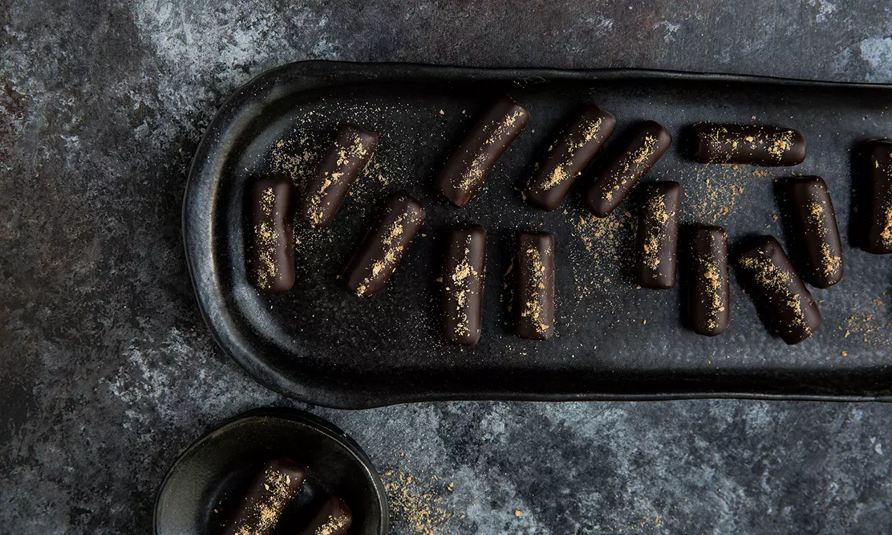 Schmale, längliche Orangen-Marzipan-Stängeli mit dunkler Schokoladenhülle auf mattschwarzer Platte, bestreut mit goldfarbenem Pulver