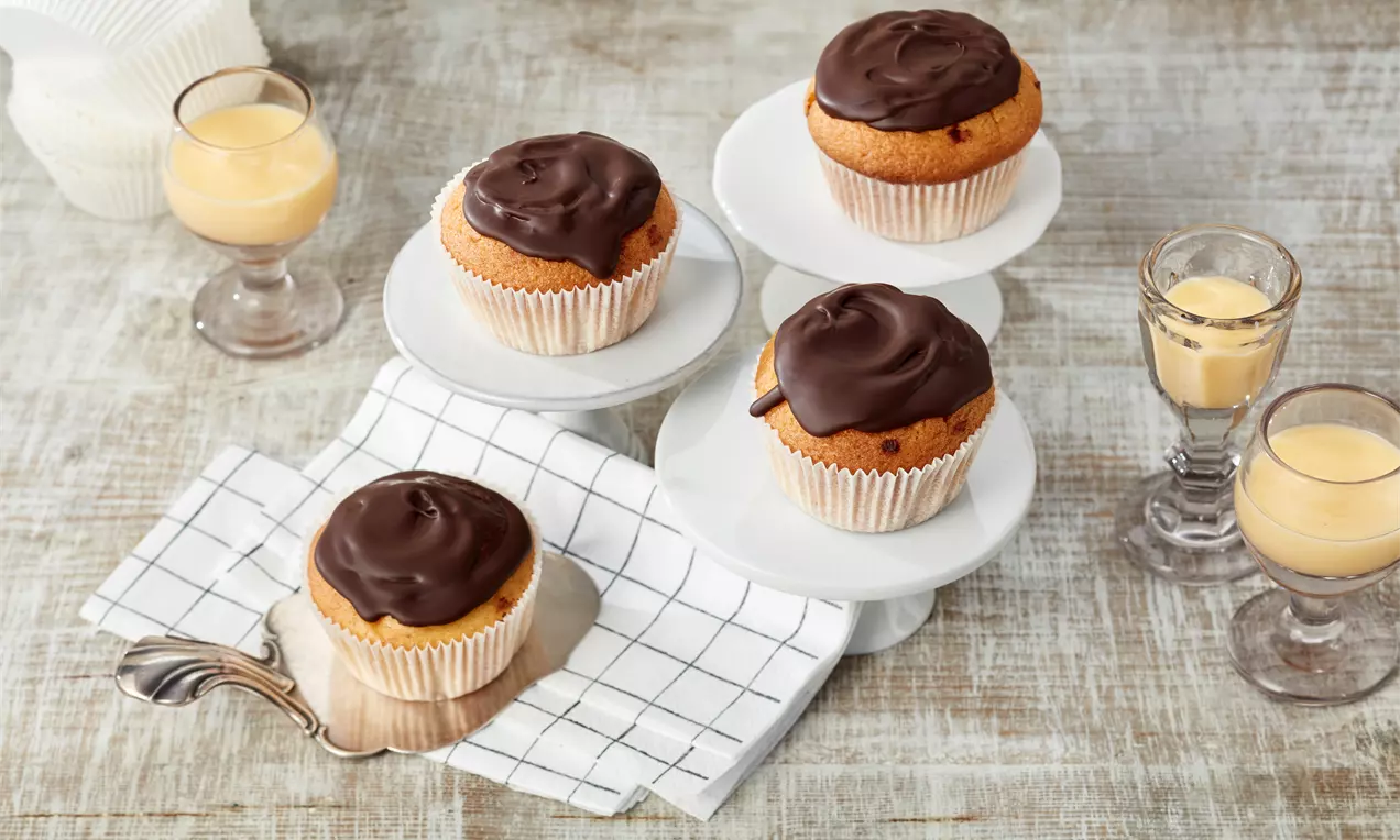 Goldbraune Eierlikör-Muffins mit glatter Schokoladenglasur in weißem Papierförmchen, auf weißen Kuchenständern und kariertem Tuch arrangiert