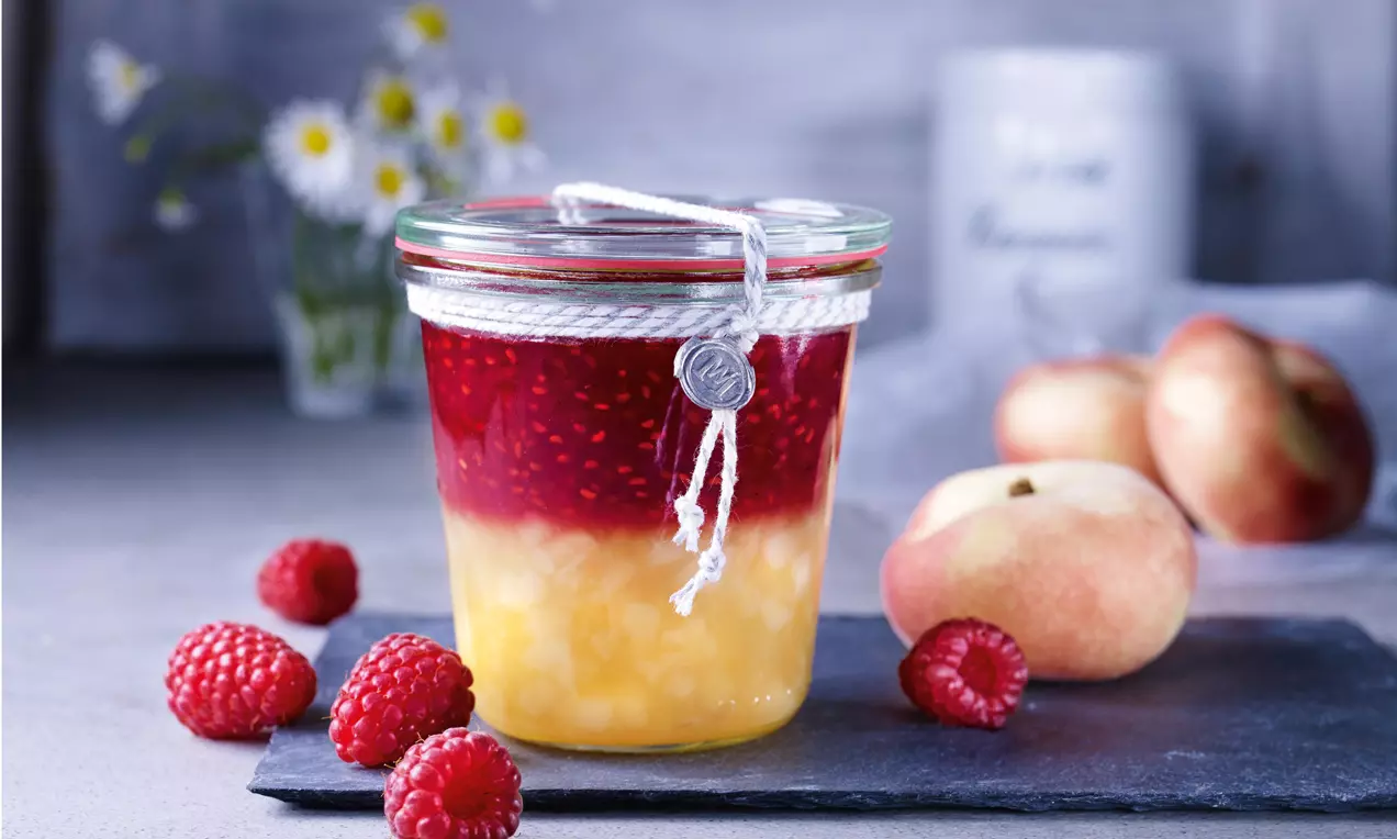 Marmellata a strati in barattolo di vetro, con base color pesca dorata e strato superiore rosso lamponi, circondato da frutti freschi su ardesia
