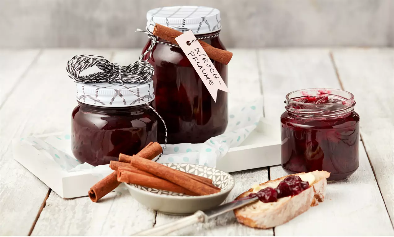 Kirsch-Pflaumen-Konfitüre in tiefroter Farbe in Gläsern mit Zimtstangen, auf Holzuntergrund mit Brot und Konfitüre auf heller Schale