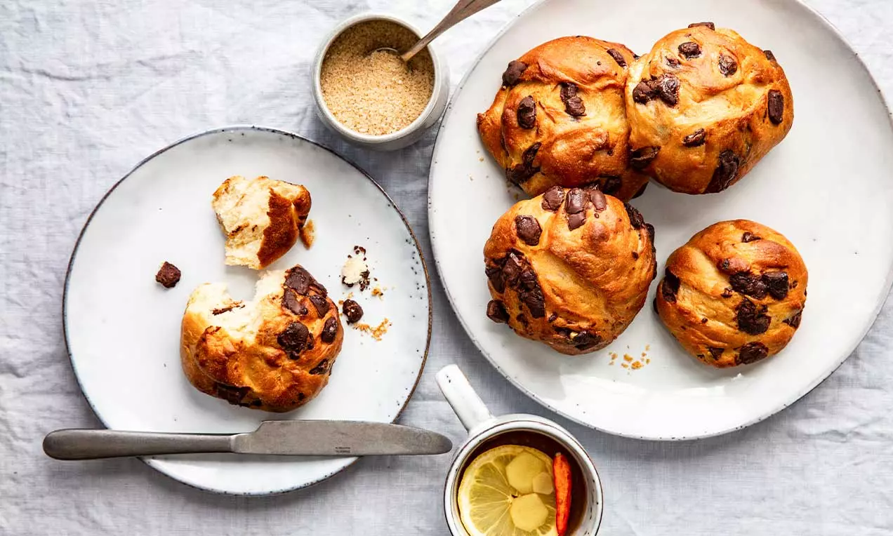 Petits pains dorés aux pépites de chocolat, légèrement brisés, disposés sur des assiettes blanches avec une tasse de thé et du sucre brun