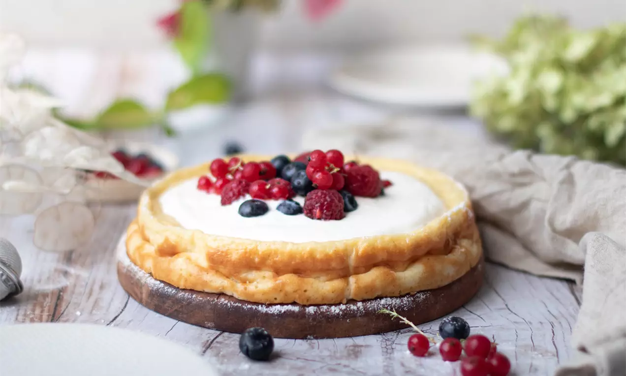 Goldbrauner Joghurtkuchen mit glatter weißer Oberfläche, belegt mit roten Johannisbeeren, Himbeeren und blauen Heidelbeeren auf hellem Holzuntergrund