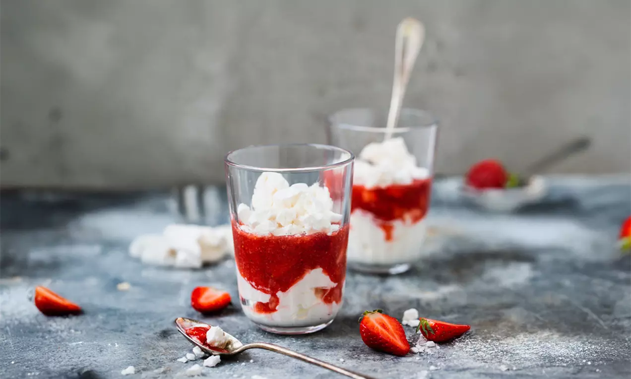 Erdbeerdessert mit leuchtend roter Fruchtschicht, heller Creme und knusprigem Baiser in gläsernen Bechern auf dunklem Tisch mit frischen Erdbeeren