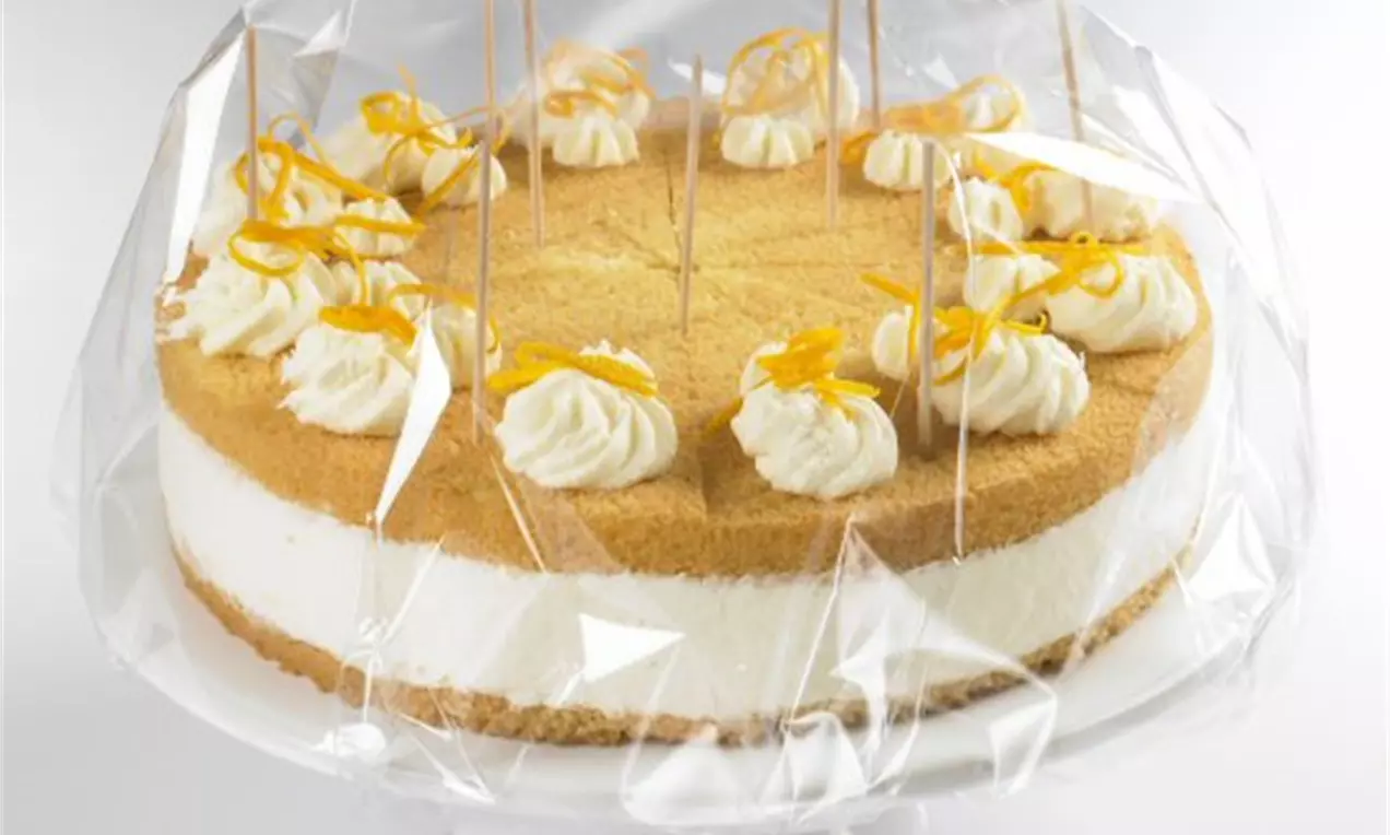 Helle, zweischichtige Torte mit goldener Oberfläche, Sahnetupfen und orangefarbenen Zesten, durch transparente Folie auf weißer Platte geschützt