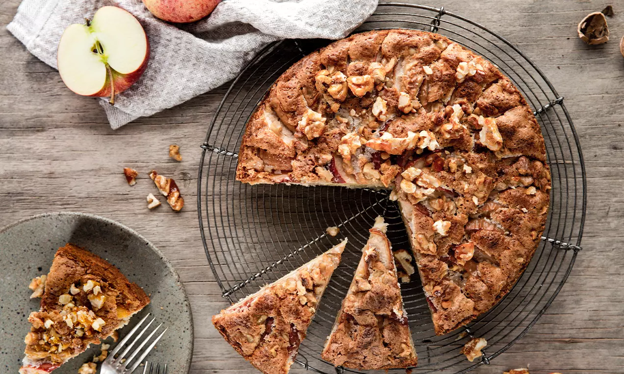 Gâteau rustique rond doré aux pommes et noix éclatées, posé sur une grille noire avec des pommes rouges et tranches sur le côté