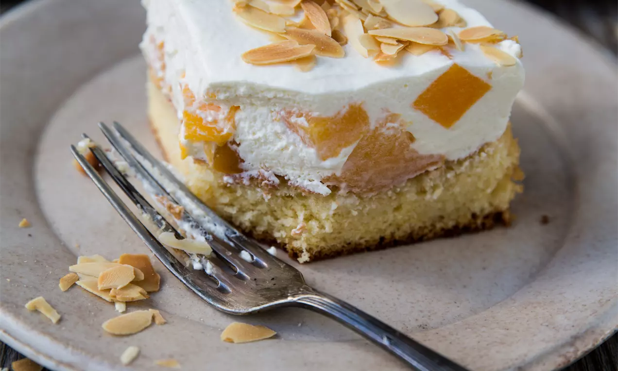 Carré de gâteau moelleux jaune doré surmonté de crème blanche, morceaux d'orange translucide et éclats d'amandes sur une assiette