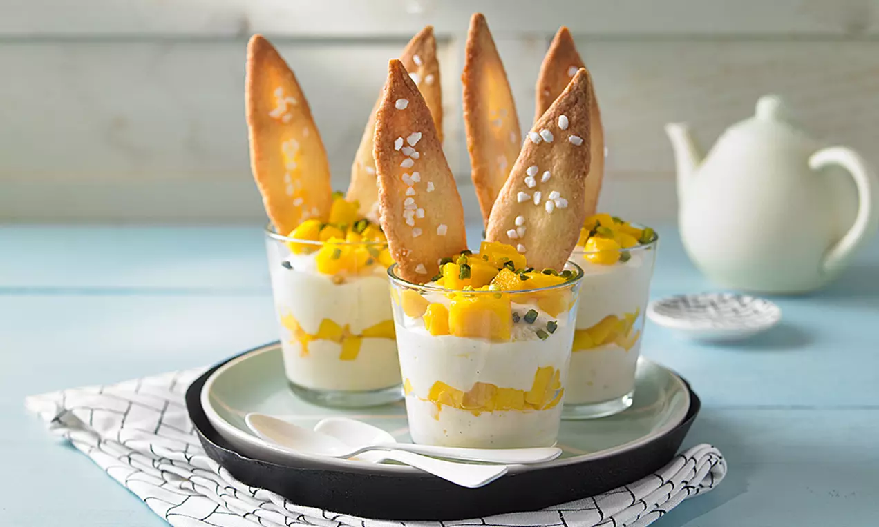 Gläser mit weißer Mango-Mousse, gelben Mangostückchen und knusprigen, gebogenen Keksöhrchen, bestreut mit grobem Zucker, auf hellem Teller