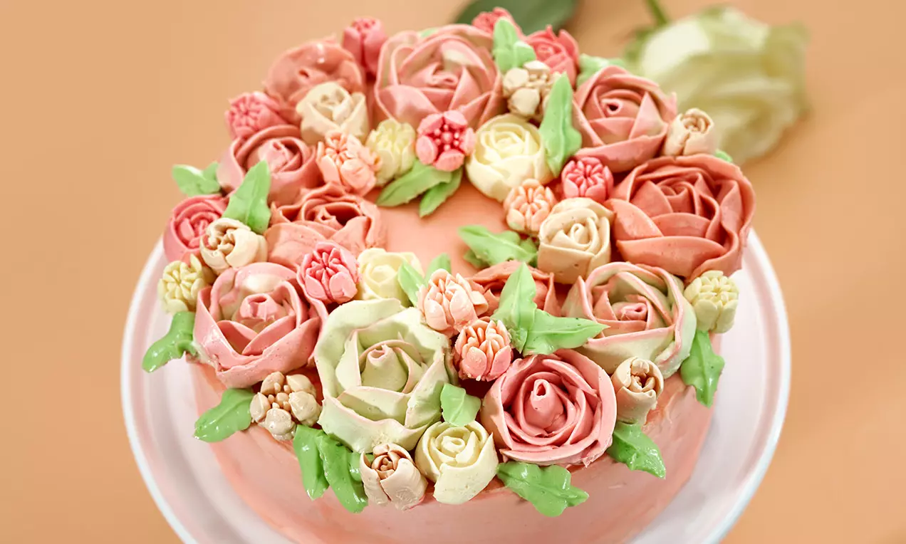 Torta rosa tenue decorata con burrosi fiori dalle sfumature crema, rosa e salmone, intervallati da foglie verdi su un piatto bianco