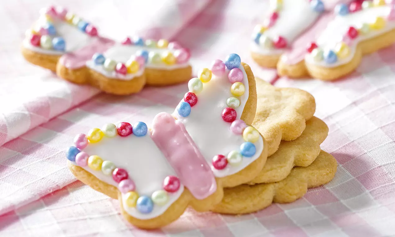 Biscotti a forma di farfalla con glassa bianca e rosa, decorati con perline colorate su una tovaglia a quadretti rosa e bianca