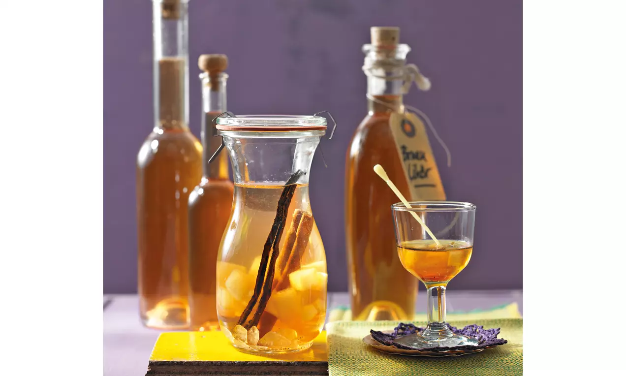 Birnenlikör in gläserner Karaffe mit Vanillestangen und Birnenstücken, daneben Likör im Glas auf lilafarbener Unterlage