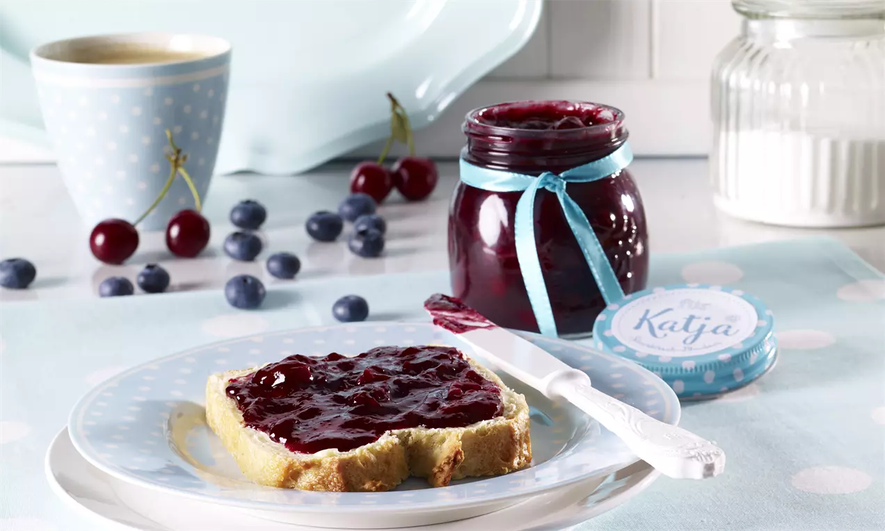 Confiture rouge vif aux cerises et myrtilles dans un pot en verre à ruban bleu, tartinée sur une tranche de pain doré avec une tasse bleue et des fruits en arrière-plan