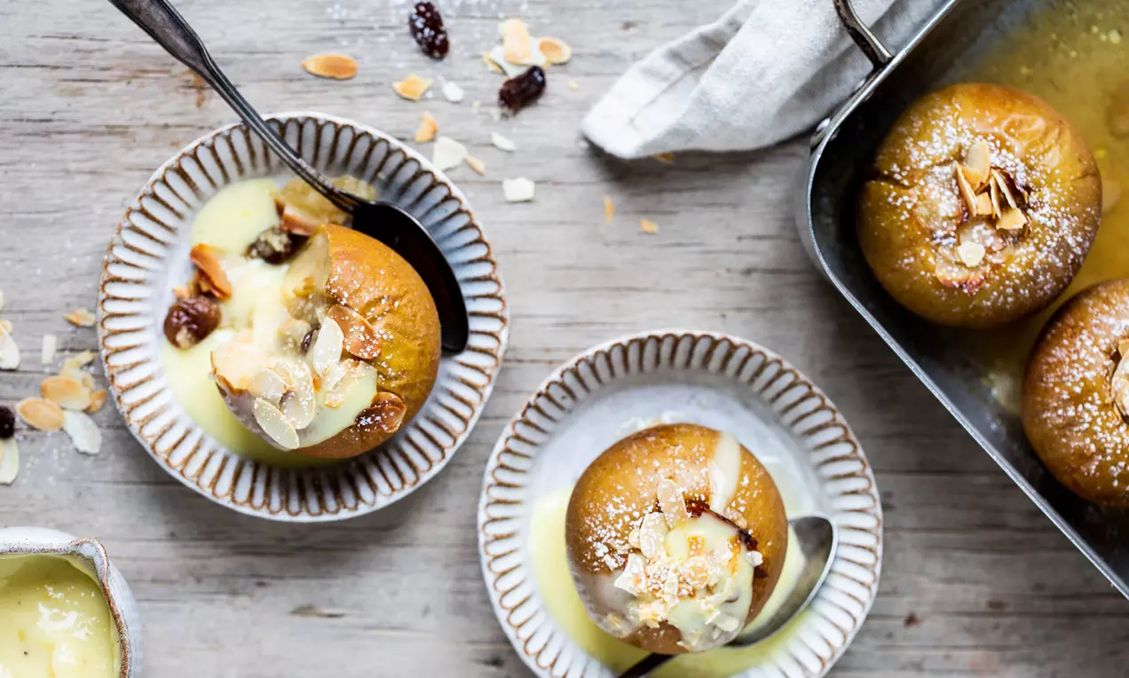 Goldbraune Bratäpfel mit geschmolzener Vanillesoße, gestreut mit Mandelblättchen und Rosinen, in rustikalen, hellen Schalen angerichtet