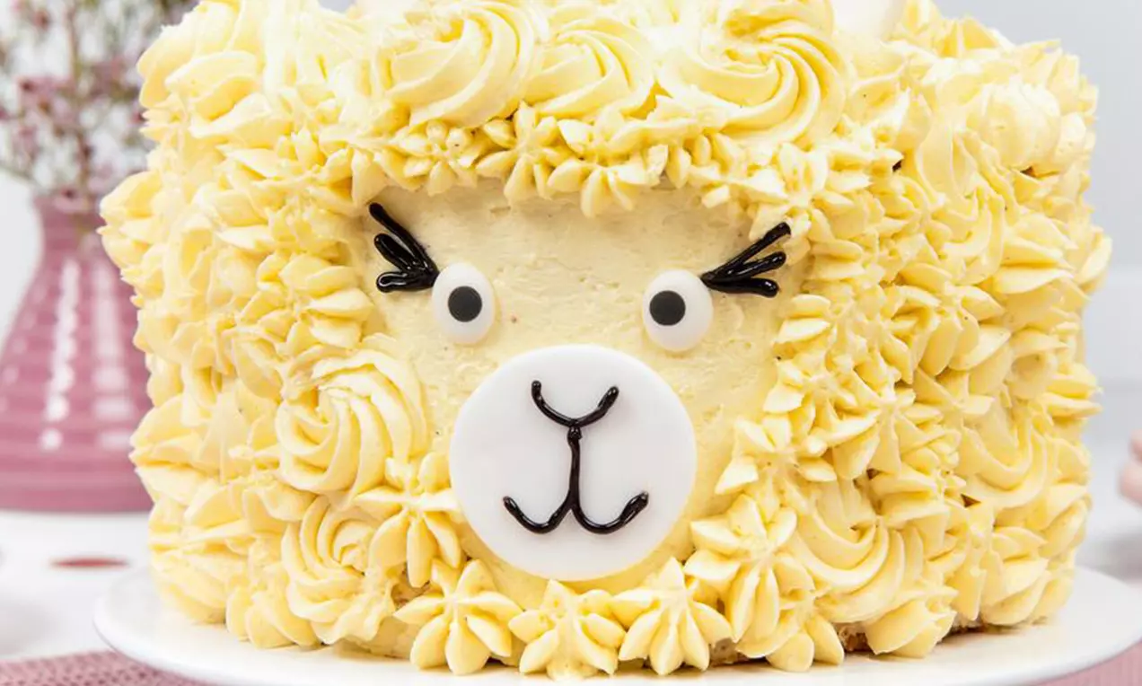 Gâteau en forme de tête de lama avec frisure de crème jaune pastel, visage en sucre blanc et noir, posé sur une assiette blanche