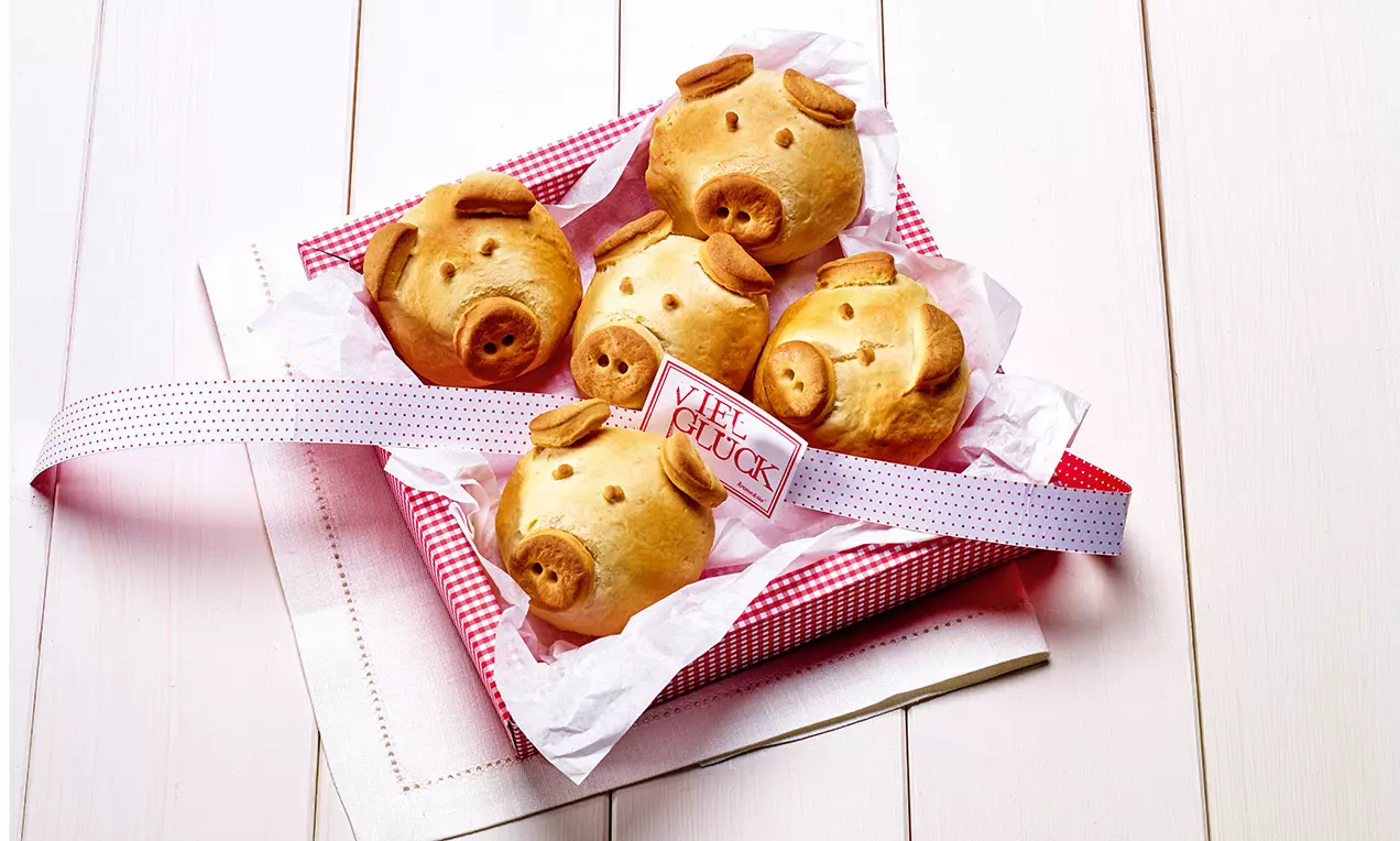 Petits pains dorés en forme de têtes de cochons avec des détails en pâte, disposés sur du papier dans une boîte rouge à carreaux blancs