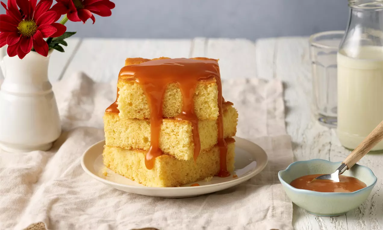 Drei helle, saftige Milchkuchenwürfel gestapelt auf einem Teller, übergossen mit glänzendem, goldbraunem Karamell, daneben eine Schale Karamellsauce