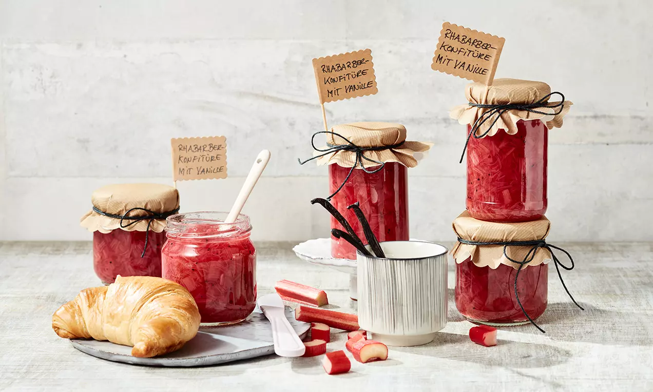 Gläser mit leuchtend roter Rhabarber-Konfitüre und beigefarbenem Papierdeckel neben Croissant, frischen Rhabarberstücken und einer weißen Tasse