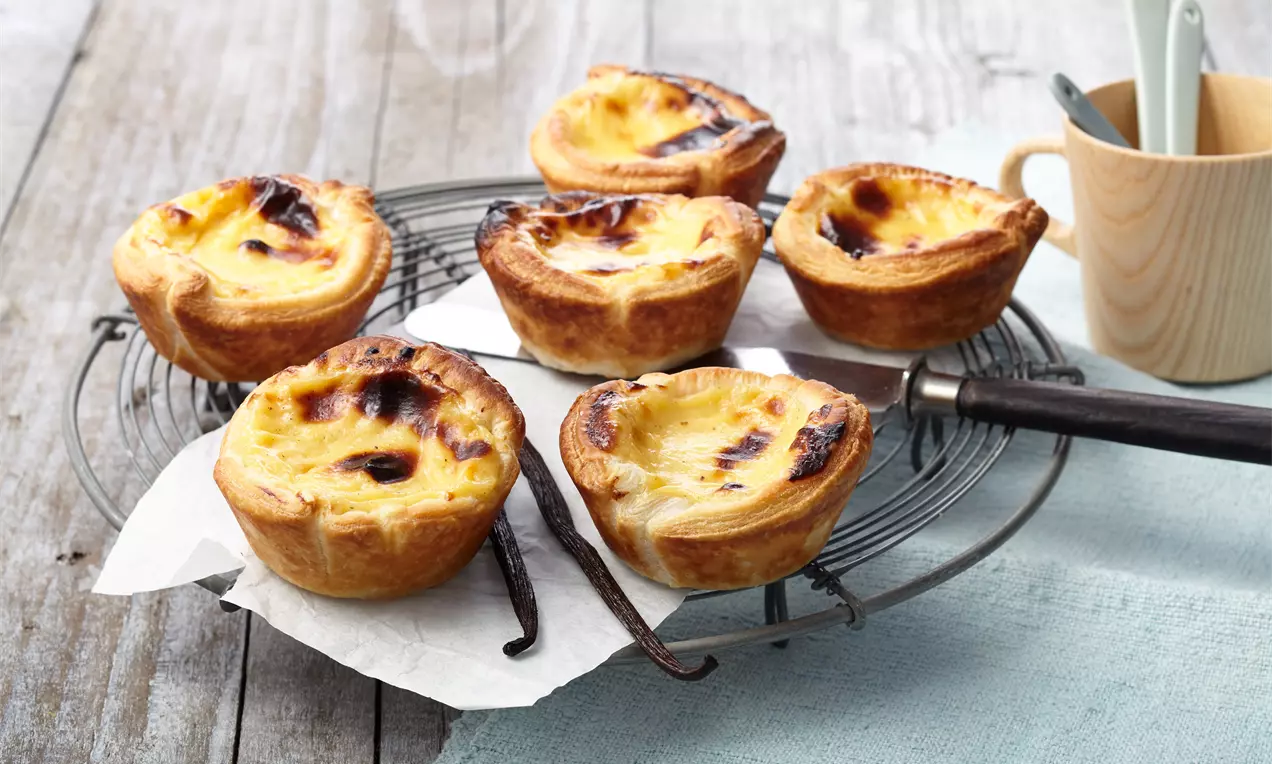 Petits flans dorés au bord croustillant dans une pâte feuilletée brune sur une grille avec des gousses de vanille et un tissu bleu
