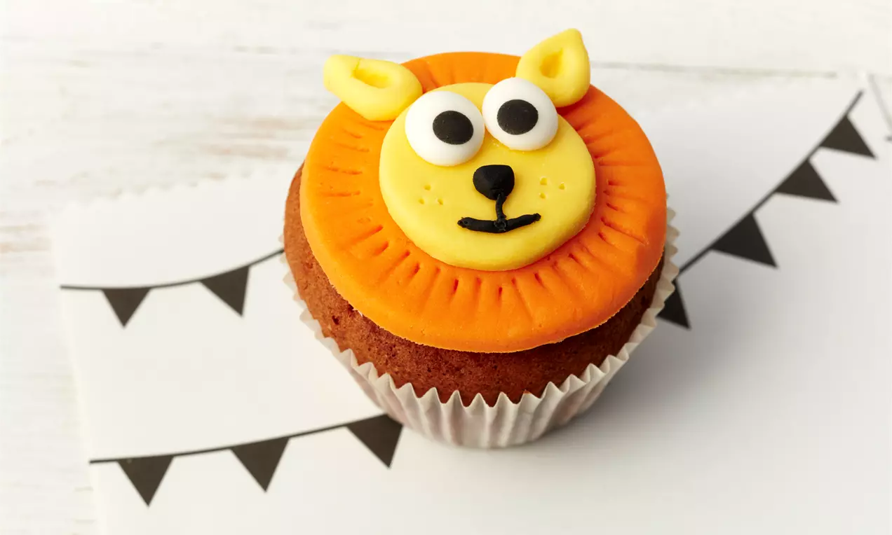 Kleiner Schokoladenmuffin in einer weißen Papierhülle, verziert mit gelbem Löwengesicht und orangefarbener Mähne aus Fondant auf einer Fahnenkarte