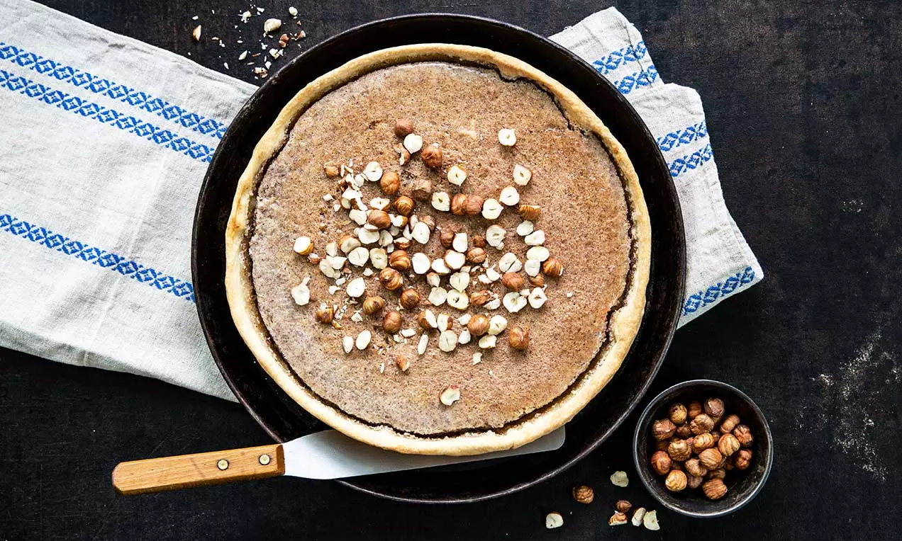 Runder Haselnusskuchen mit goldbraunem Rand und cremiger Oberfläche, bestreut mit gehackten Haselnüssen in einer schwarzen Backform auf einem Tuch