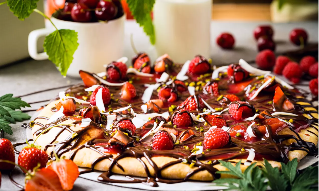Pizza sucrée grillée dorée garnie de fraises fraîches, framboises, chocolat fondu et copeaux blancs sur une table entourée de fruits rouges