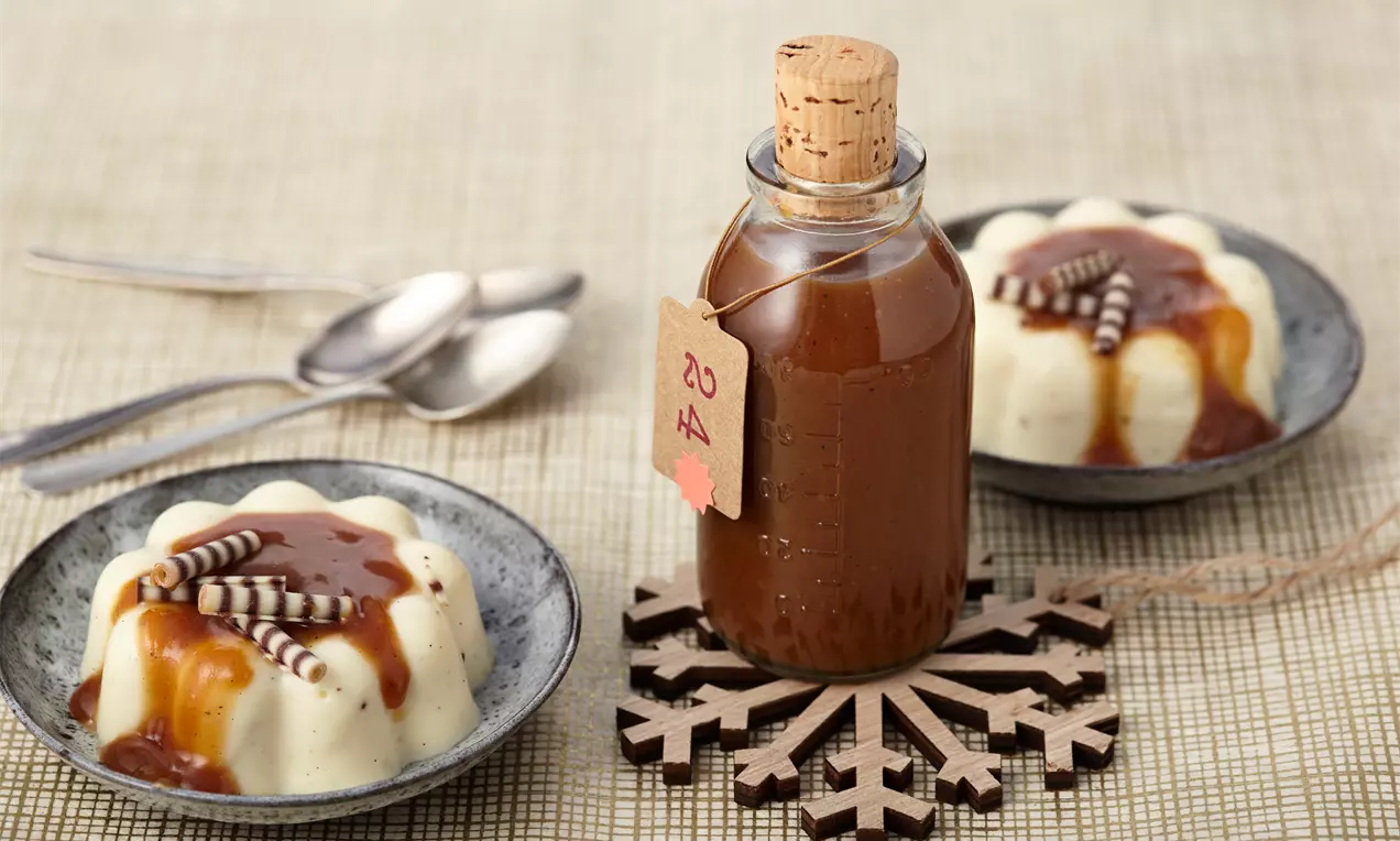 Braune Toffeesoße in einer kleinen Glasflasche mit Korkverschluss, arrangiert auf einem Holz-Schneeflocken-Untersetzer neben zwei Puddingdesserts
