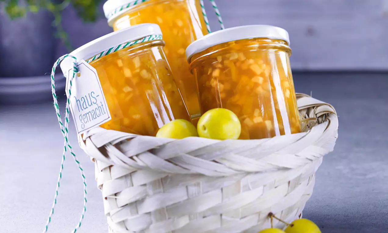 Drei Gläser mit goldgelber Apfel-Mirabellen-Konfitüre in einem weiß geflochtenen Korb, dekoriert mit gelben Mirabellen vor grauem Hintergrund