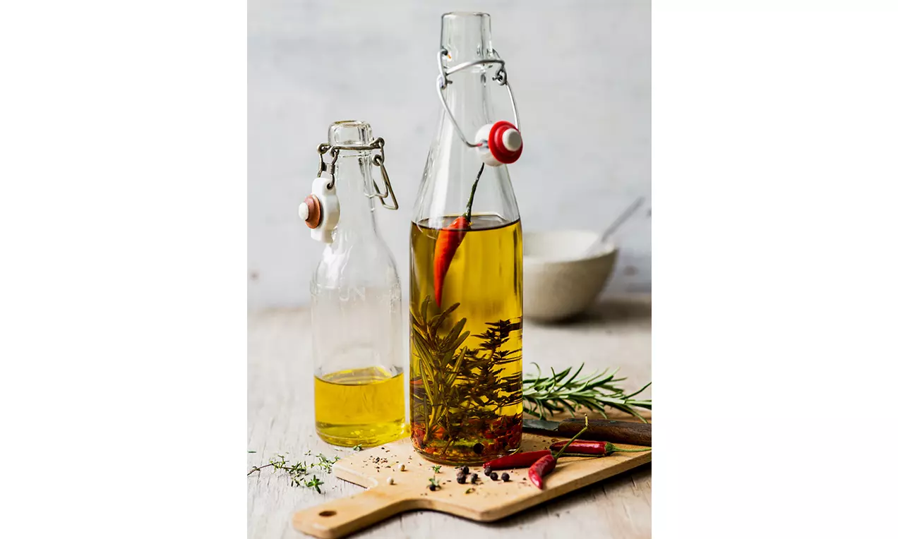 Goldgelbes Peperoncini-Öl in klarer Glasflasche mit roten Chilischoten, Rosmarinzweigen und Pfefferkörnern auf hölzernem Schneidebrett