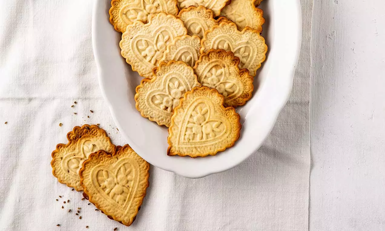 Biscotti Züri-Tirggel dorati a forma di cuore con bordi ondulati e motivi intricate sulla superficie, disposti in un piatto bianco ovale su una tovaglia candida