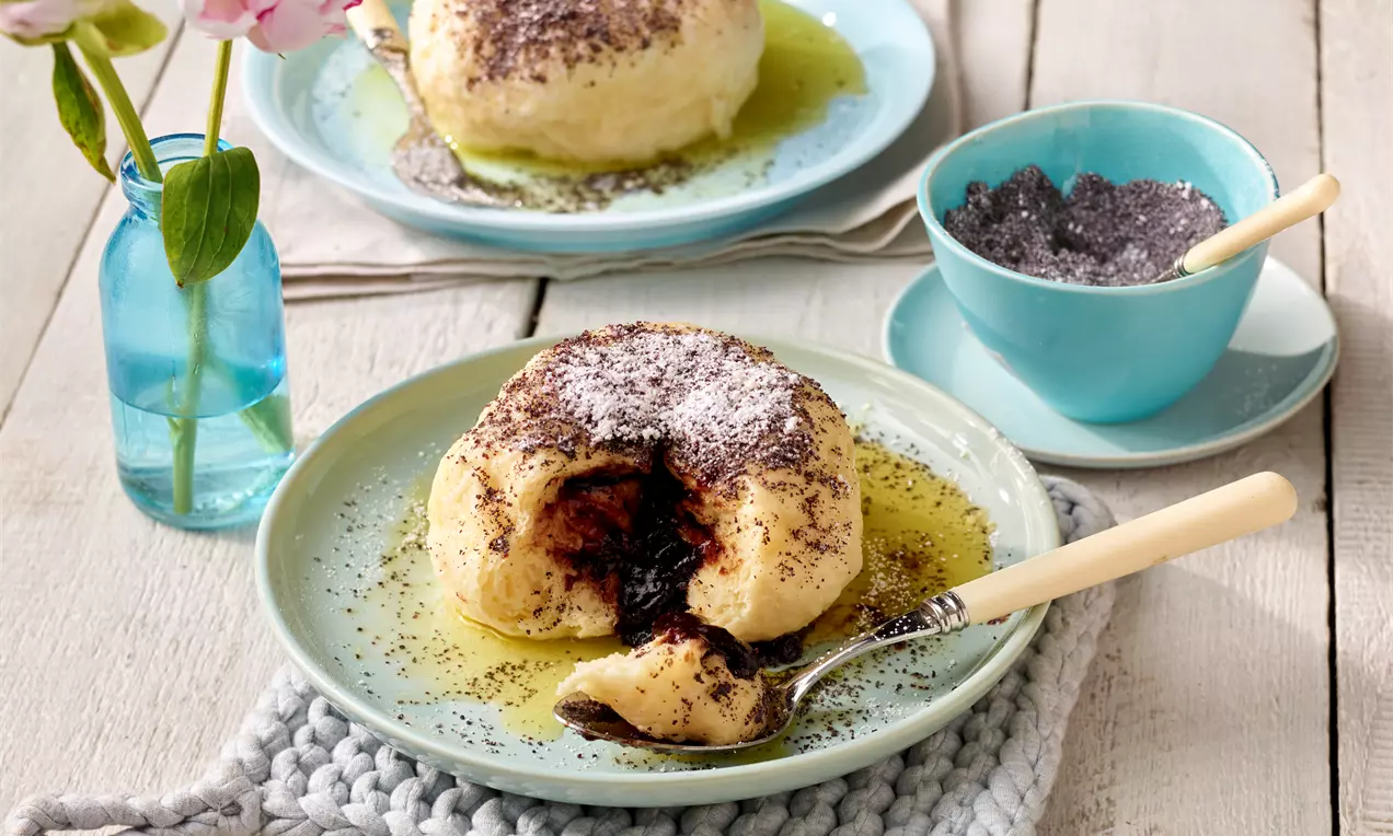 Germknödel mit hellem Teig, gefüllt mit Pflaumenmus, bestreut mit Mohn und Puderzucker, serviert auf hellblauem Teller mit zerlaufener Butter