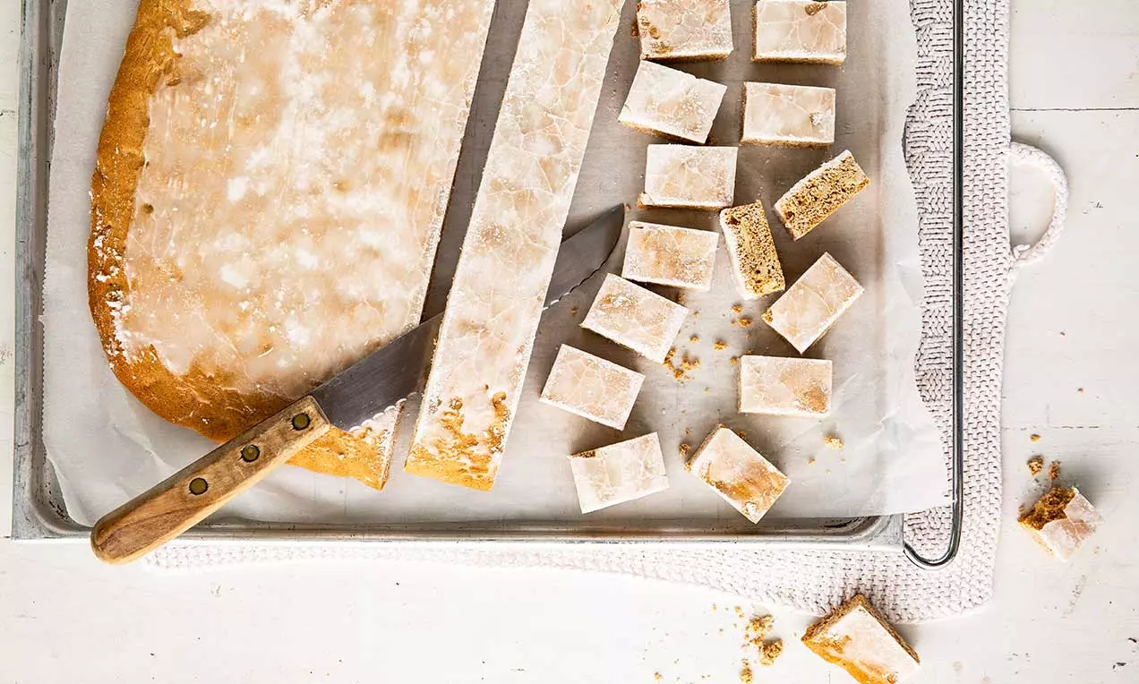 Rechteckige Basler Läckerli mit glasierter Oberfläche in hellem Beige, auf Backpapier geschnitten und in Würfeln locker verteilt, daneben ein Messer