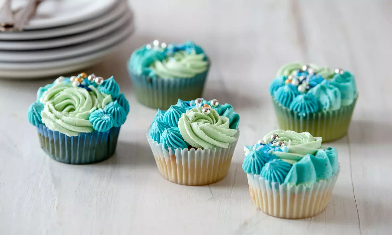 Cupcakes mit türkis- und mintfarbener Buttercremeblüten, verziert mit silbern und golden schimmernden Perlchen auf hellem Holztisch
