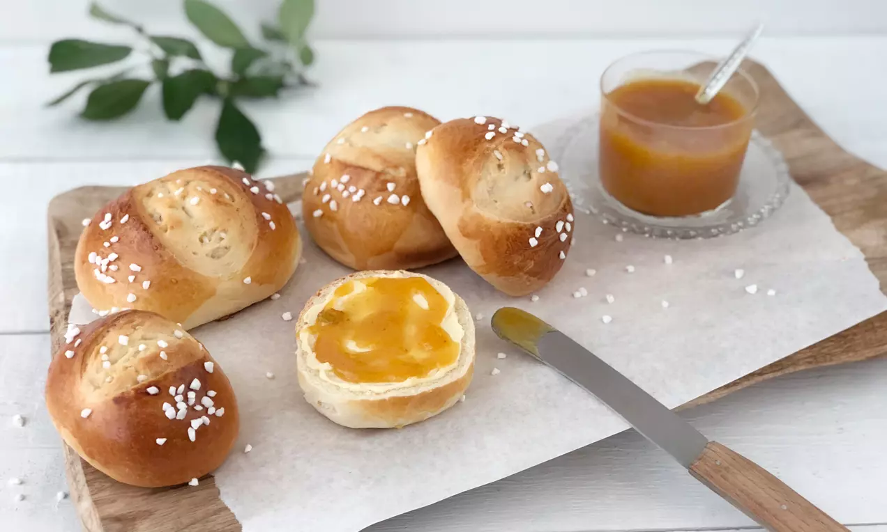 Goldbraune, rund geformte süße Brötchen mit Hagelzucker auf weißem Papier, ein aufgeschnittenes Brötchen mit Marmelade neben Glas und Messer
