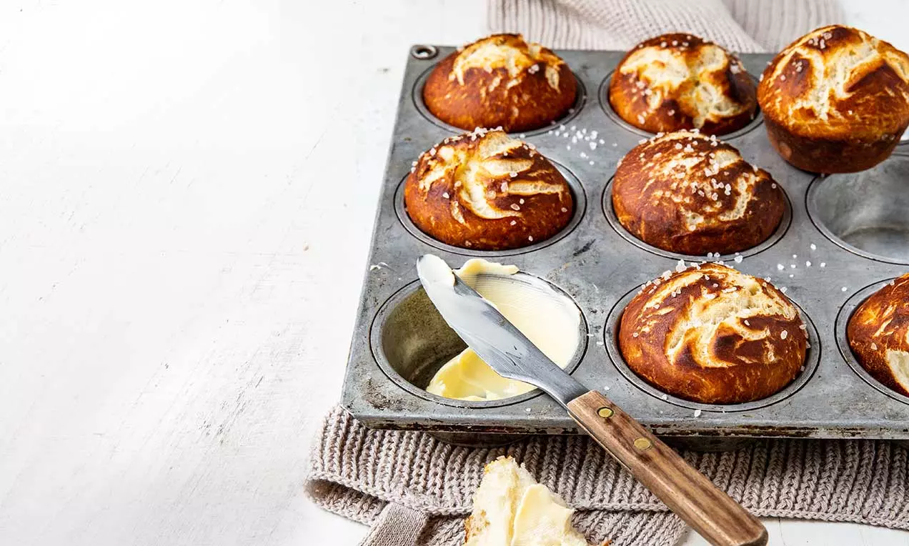 Goldbraune, rundliche Laugenbrötli mit weißem Salz bestreut, in einer Metallform gebacken, daneben Butter mit Messer auf Stofftuch drapiert