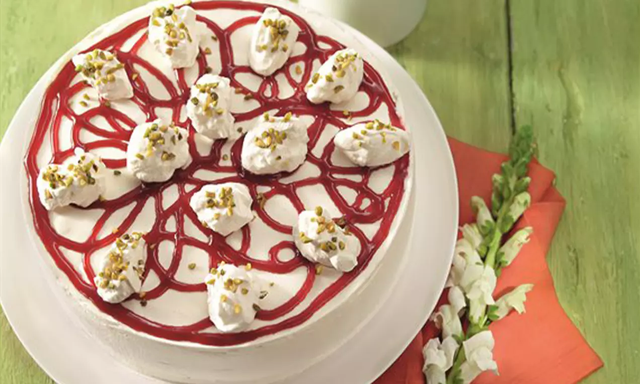 Holunderblüten-Torte mit weißer Cremeschicht, roten Sektglasuren, Sahnetupfen und Pistaziensplittern auf einer weißen Platte auf grünem Untergrund