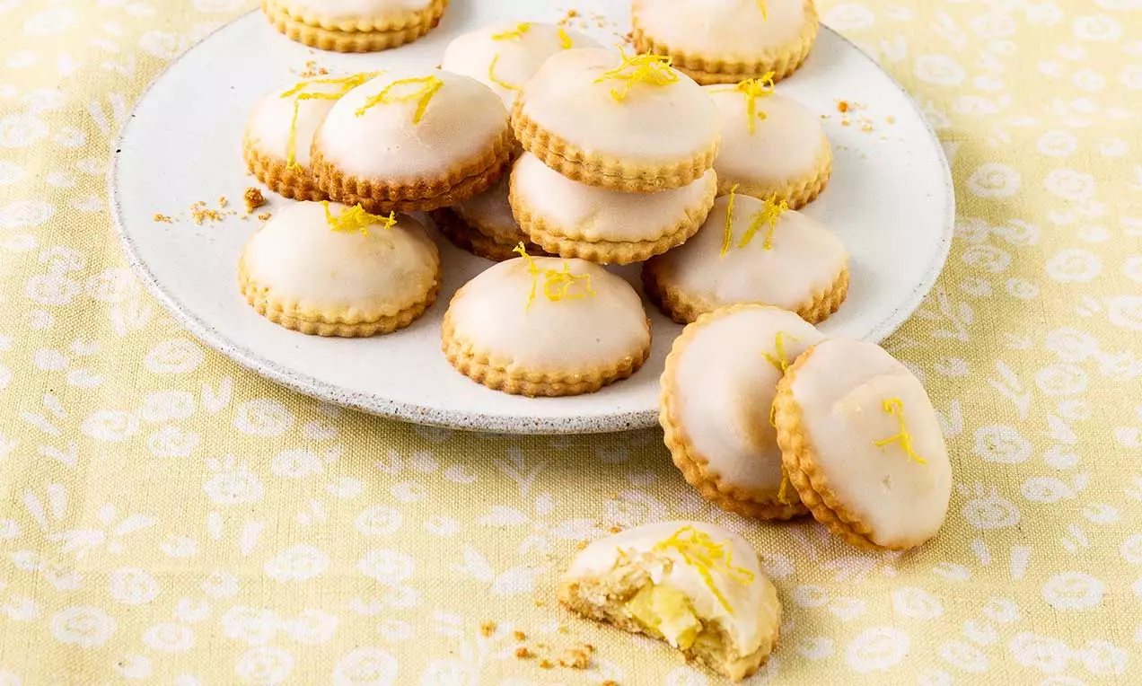 Biscotti rotondi e dorati con bordo ondulato, glassa chiara e scorza di limone, disposti su un piatto bianco su tovaglia gialla decorata