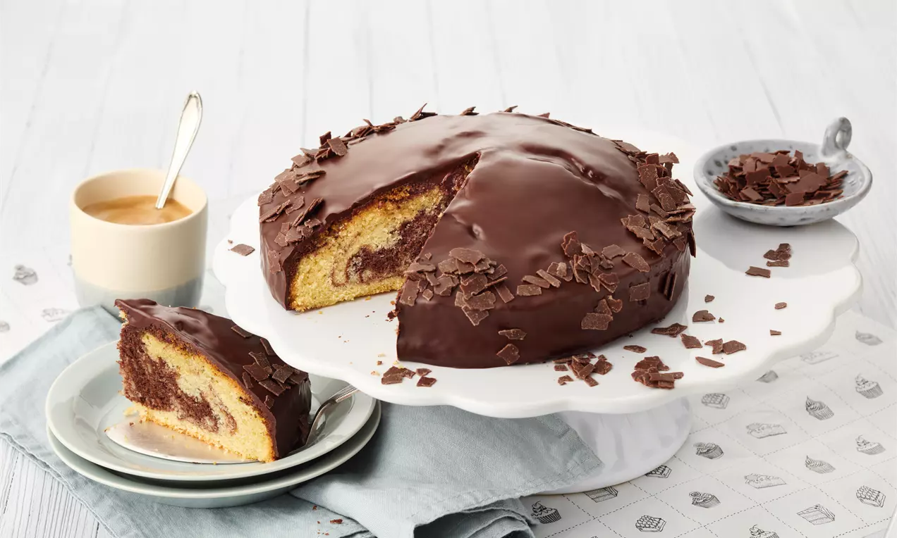 Gâteau marbré moelleux aux nuances de chocolat et vanille, enrobé de glaçage brillant au chocolat et décoré de copeaux sur un plat blanc