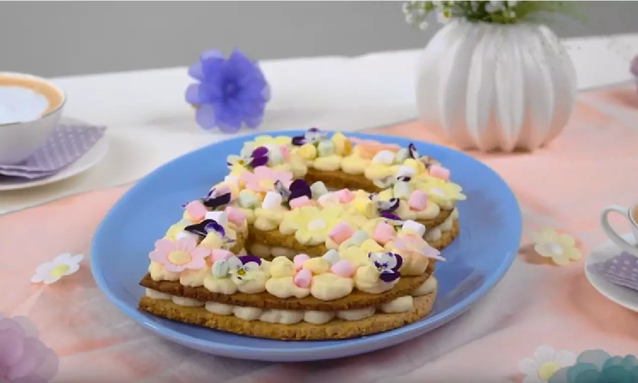 Gâteau lettre en pâte sablée dorée garnie de crème blanche, fleurs pastel en sucre et violettes sur une assiette bleue