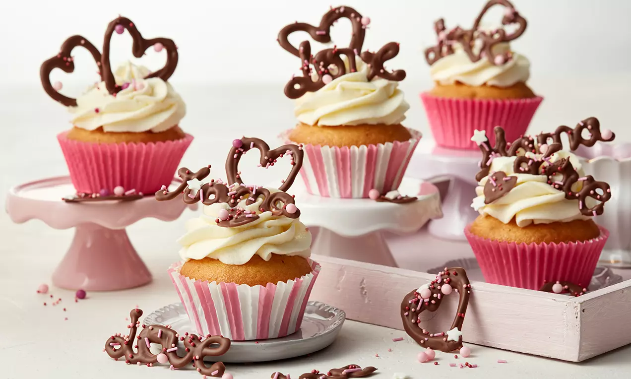 Vanillefarbene Cupcakes mit weißer Cremetopping, rosa Papierförmchen und filigraner Schokoladendekoration auf dekorativem rosa Geschirr