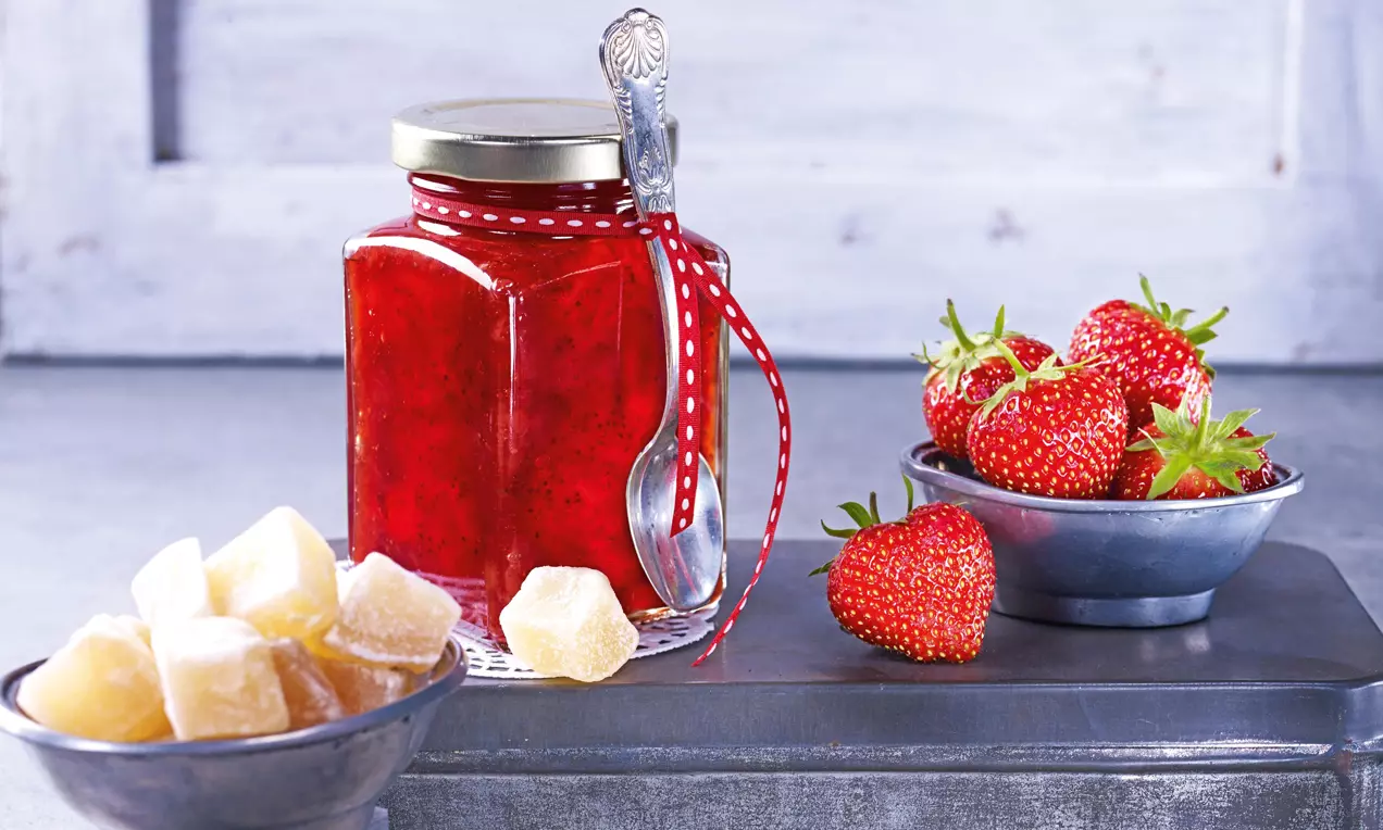 Glas mit tiefroter Erdbeer-Ingwer-Konfitüre, verziert mit rotem Band, daneben frische Erdbeeren und kandierter Ingwer in Schalen auf grauem Untergrund
