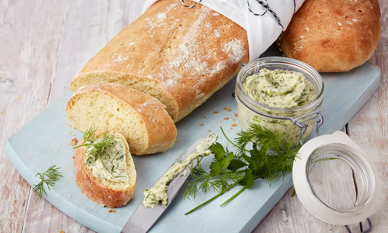 Goldbraunes Ciabatta mit knuspriger Kruste, angeschnitten auf hellblauem Holzbrett, daneben cremige Kräuterbutter in Glas und frische Kräuter