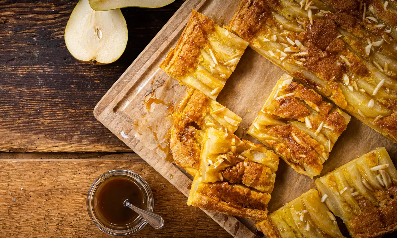 Gebräunte Birnen-Galette-Stücke mit goldgelber Oberfläche, Mandelsplittern und Karamellsoße auf Holzbrett neben angeschnittenen Birnen