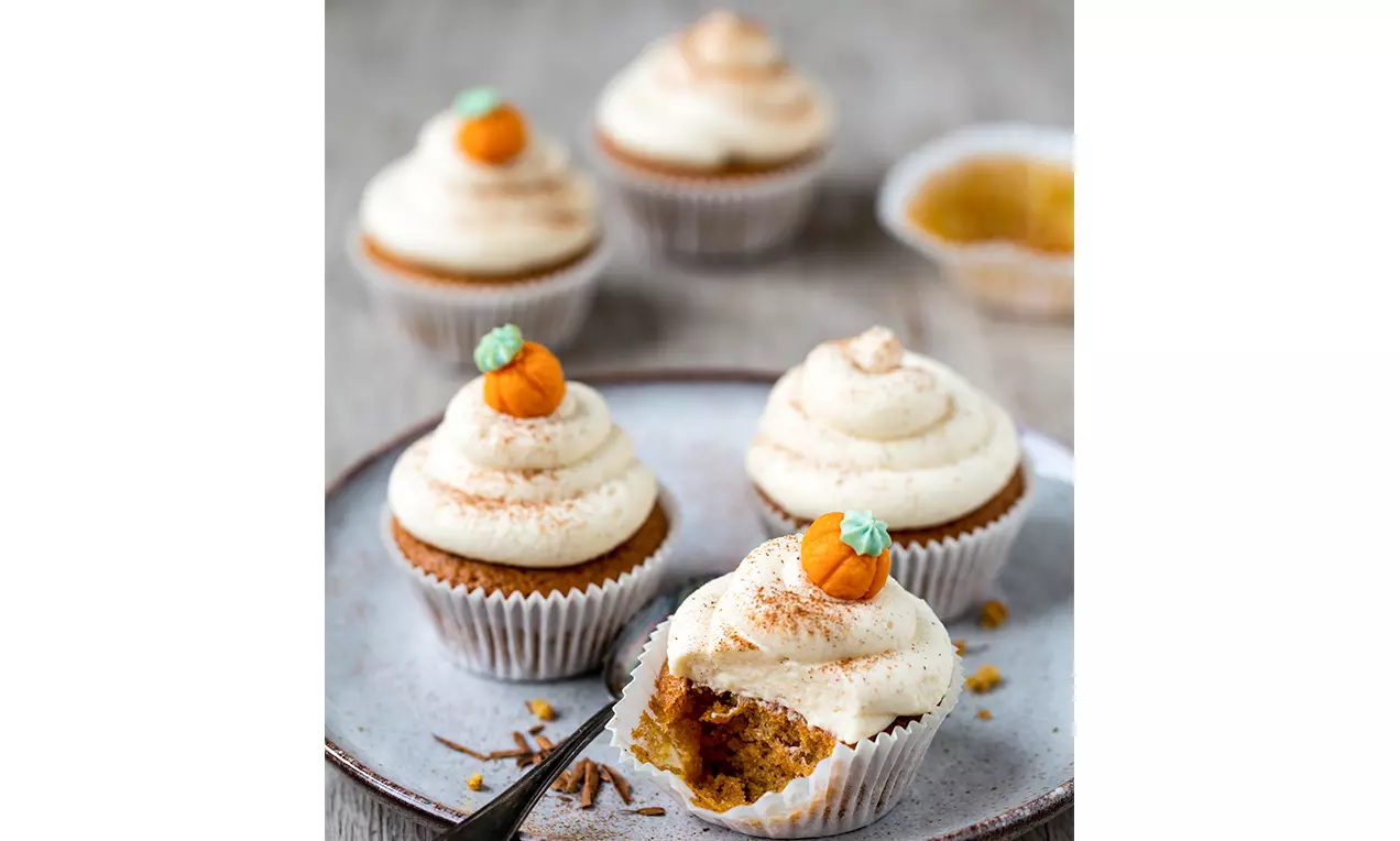 Cupcake color caramello con glassa bianca cremosa a spirale, decorato con una piccola zucca arancione e servito su un piatto chiaro
