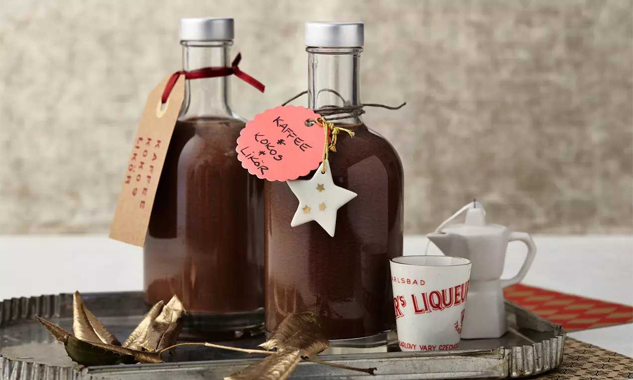 Zwei klare Glasflaschen mit dunkelbraunem Kaffee-Kokos-Likör, dekoriert mit Anhängern, auf silbernem Tablett mit Goldsternen und weißem Porzellangefäß