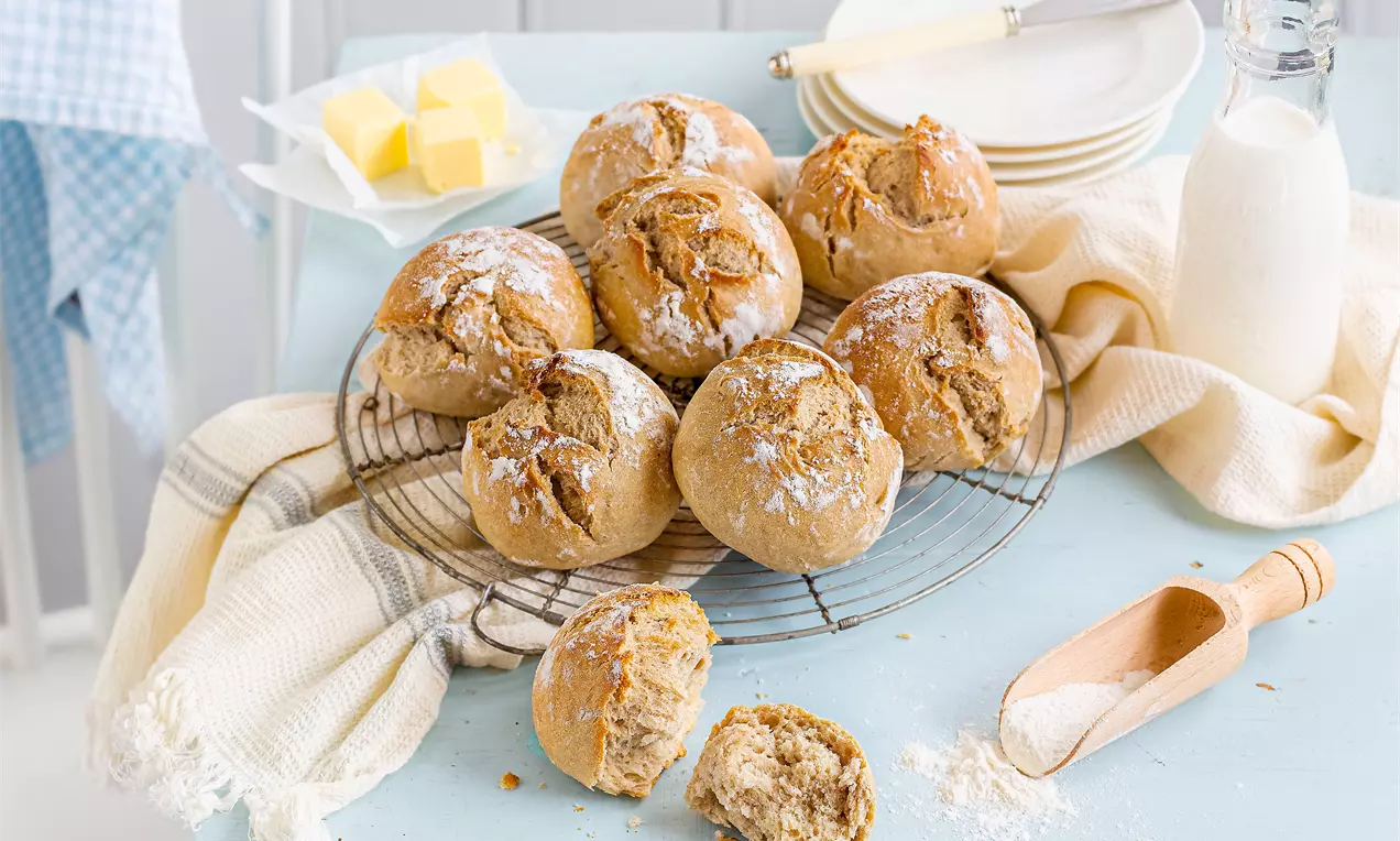 Goldbraune Sauerteig-Brötchen mit rissiger Kruste auf einem runden Rost, umgeben von Butterwürfeln, einer Milchflasche und Mehl auf hellem Untergrund