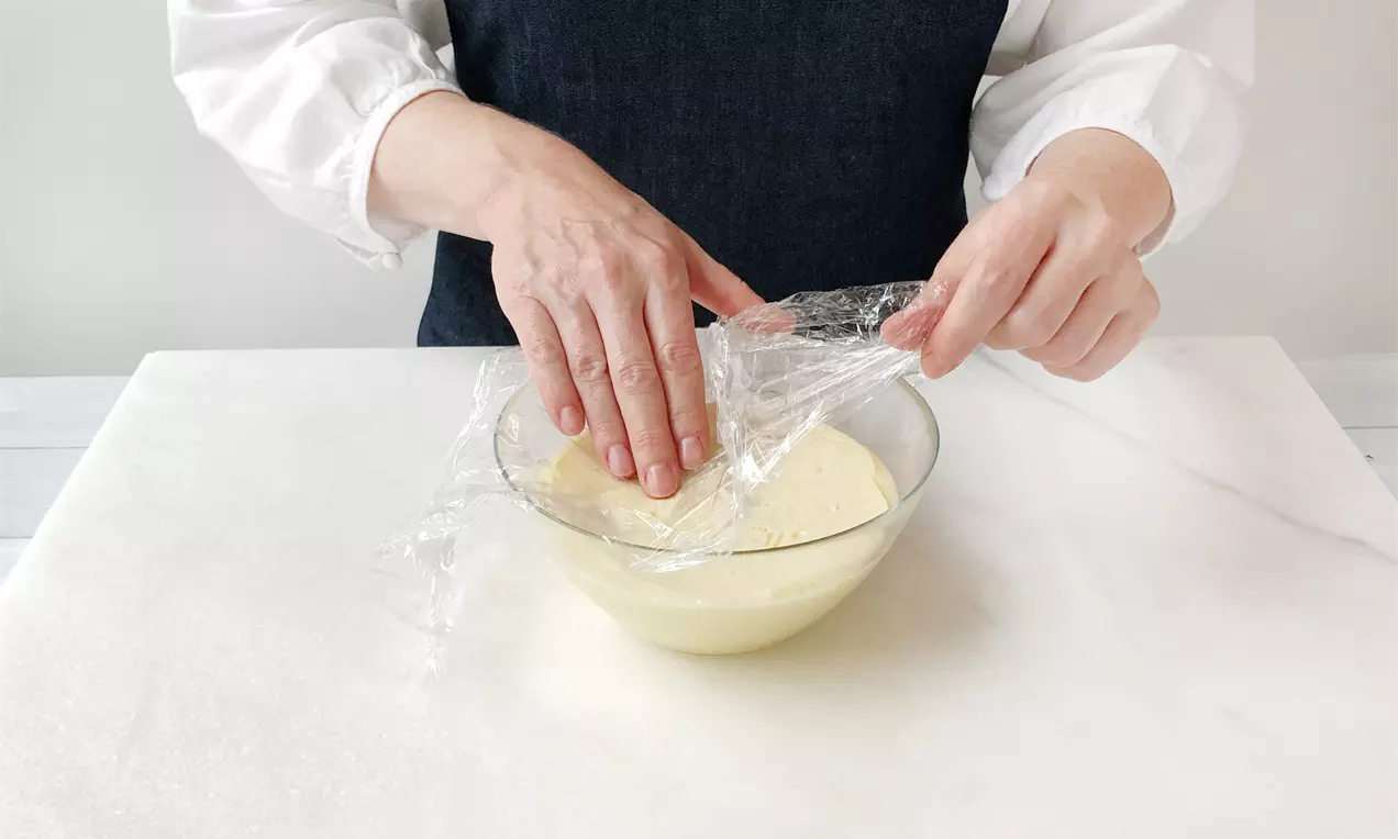 Hand zieht transparente Frischhaltefolie über eine glatte, helle Puddingoberfläche in einer klaren Glasschüssel auf weißer Arbeitsfläche