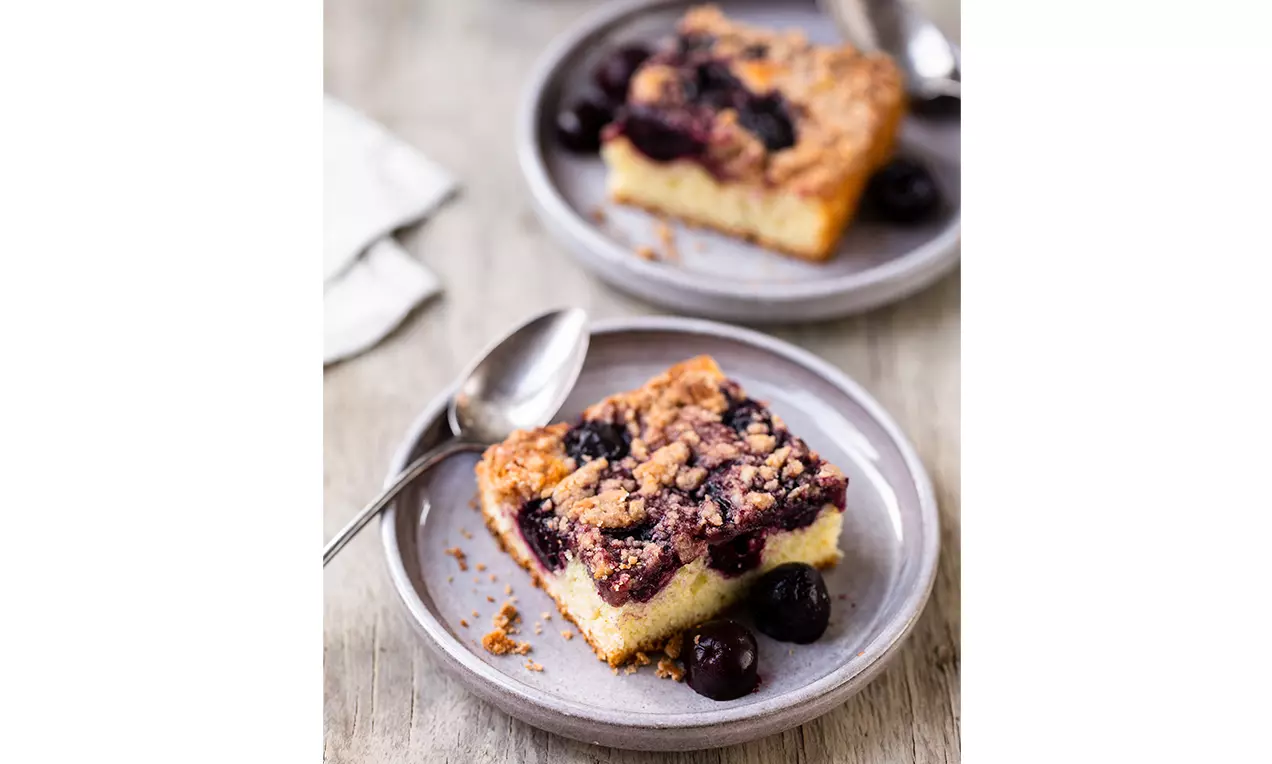 Carré moelleux à la pâte dorée surmonté de cerises juteuses et d’un crumble croustillant servi sur une assiette grise avec une cuillère