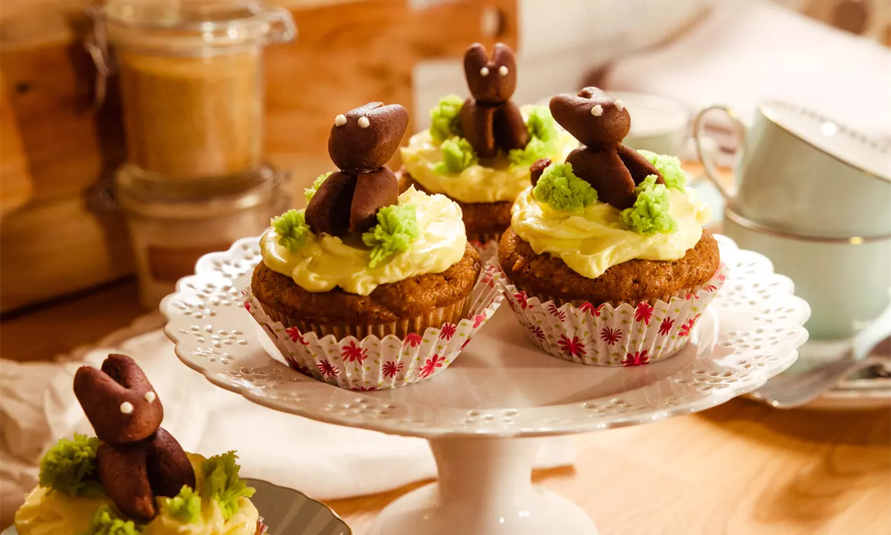Vegane Möhren-Nuss-Cupcakes in gemusterten Papierförmchen mit heller Creme, grüner Deko und schokoladenbraunen Hasenfiguren auf weißer Tortenplatte