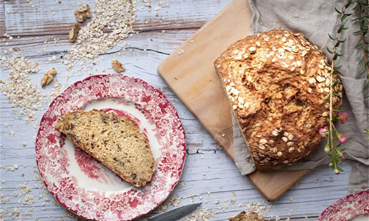 Brot mit Haferflocken als Deko, das auf einem beigen Tuch liegt, mit einem Porzellanteller und Ästen, die im Hintergrund sichtbar sind.